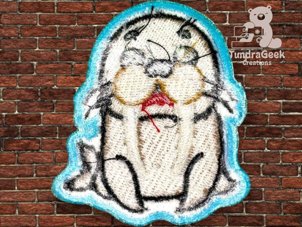 An embroidered patch featuring a cartoon seal with red thread near its mouth, displayed against a brick wall background. TundraGeek Creations logo appears in the top right corner.