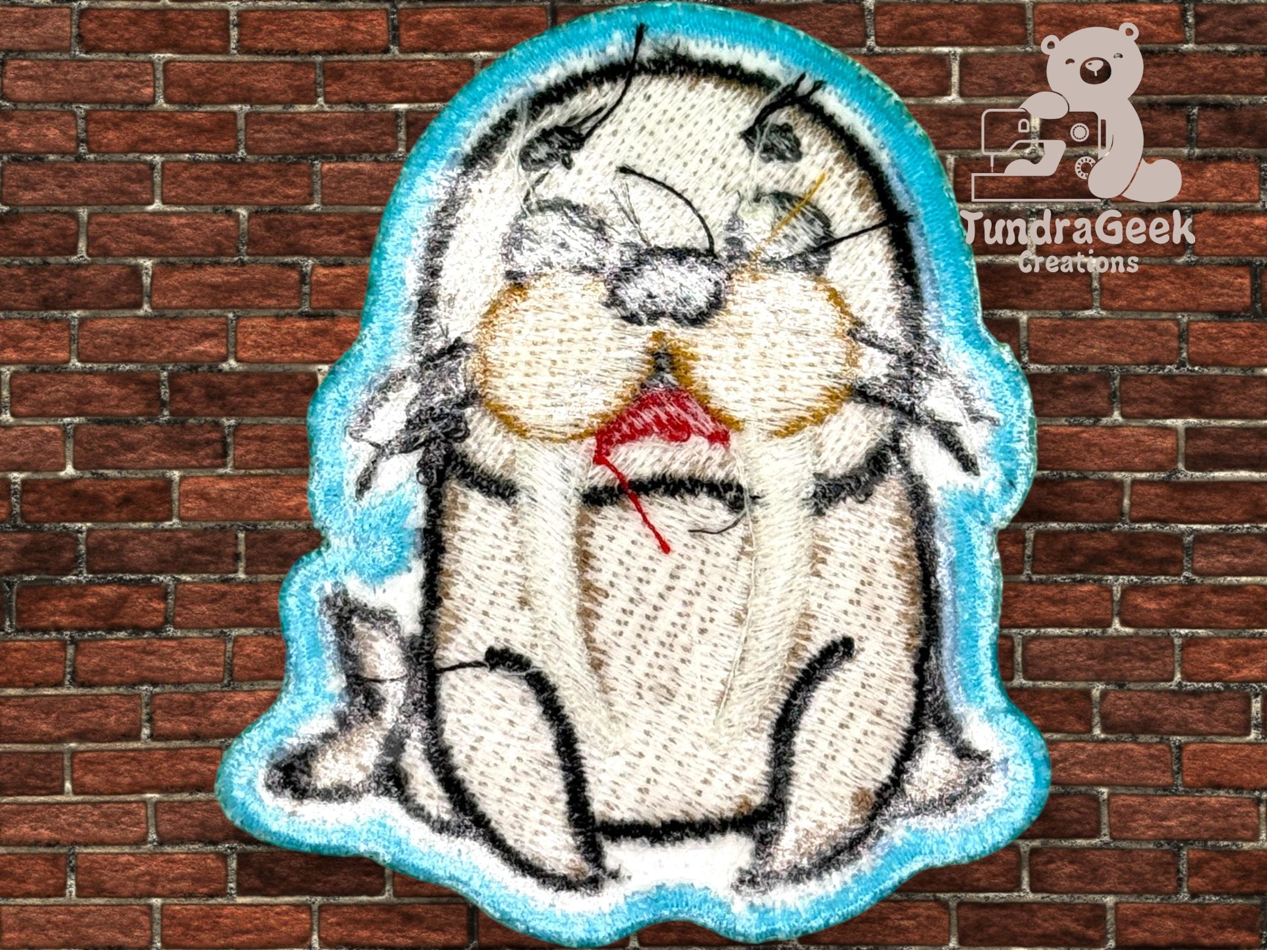 An embroidered patch featuring a cartoon seal with red thread near its mouth, displayed against a brick wall background. TundraGeek Creations logo appears in the top right corner.