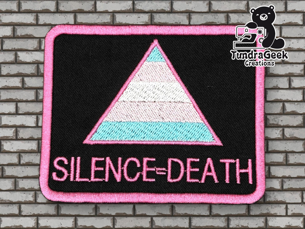 Rectangular patch with a pink-bordered triangle in trans pride colors and the words SILENCE=DEATH below, on a black background. Brick wall backdrop, TundraGeek Creations logo in corner.