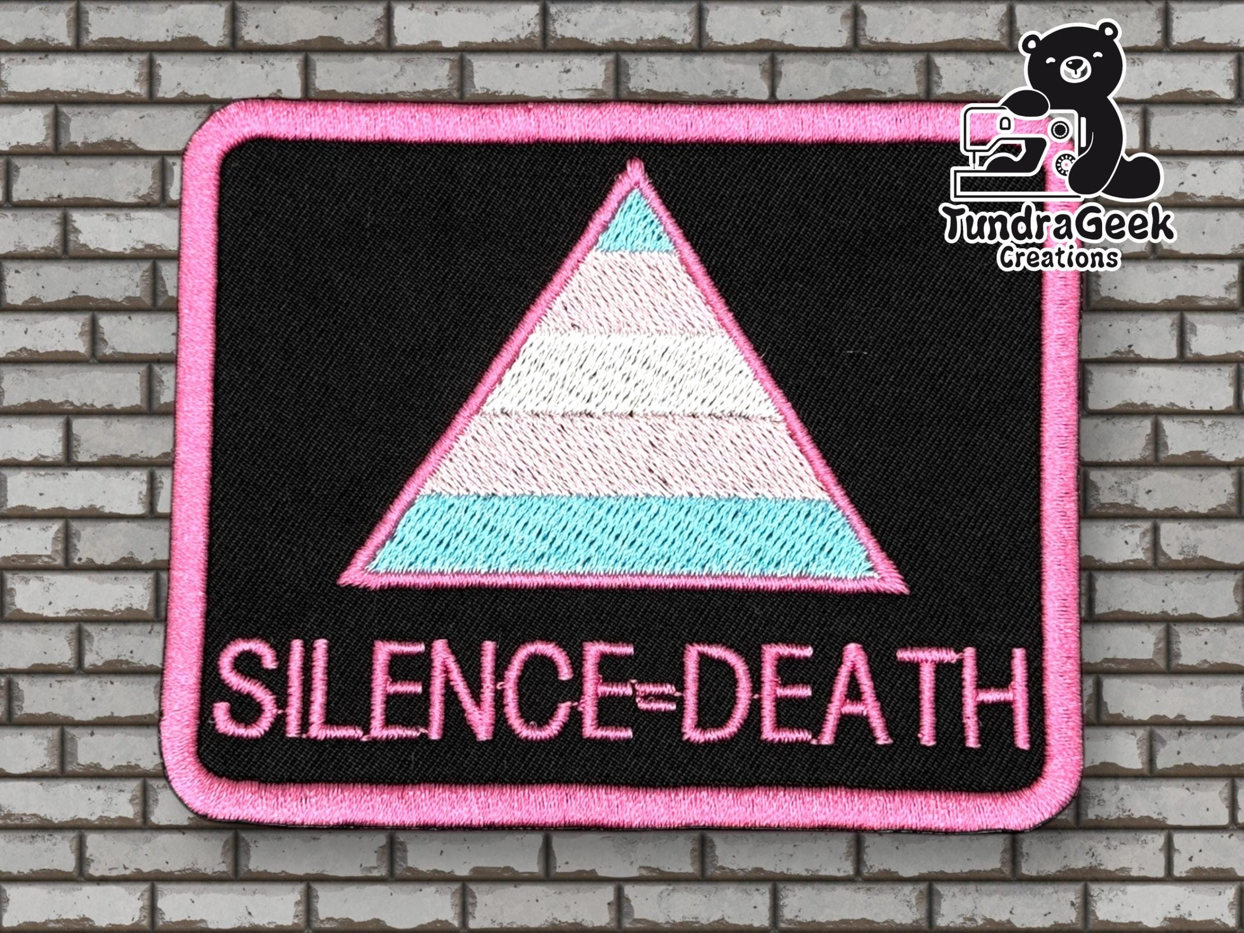 Rectangular patch with a pink-bordered triangle in trans pride colors and the words SILENCE=DEATH below, on a black background. Brick wall backdrop, TundraGeek Creations logo in corner.