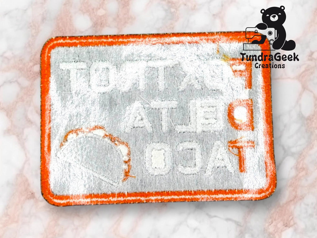 An embroidered patch with reversed text TACOT DELTA TACT outlined in orange, dusted with white powder, on a marble surface. TundraGeek Creations logo in the top right corner.
