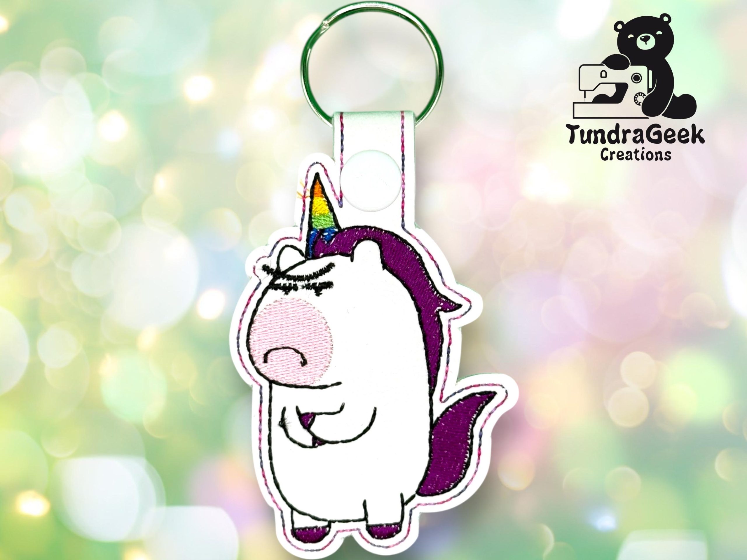 Embroidered keychain featuring a grumpy unicorn with a rainbow horn and purple mane, displayed on a blurred pastel background. TundraGeek Creations logo in the corner.