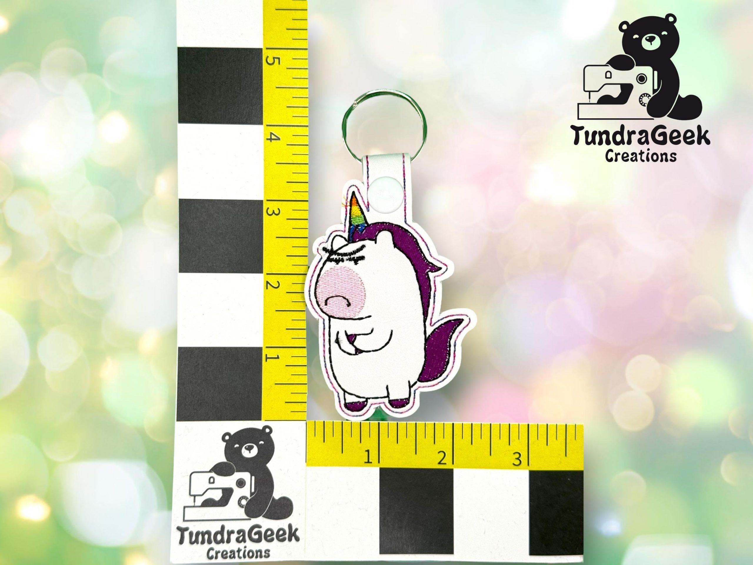 A keychain featuring a cartoon unicorn character is shown next to rulers for scale, measuring approximately 3 inches in height. TundraGeek Creations logo is visible in two places.