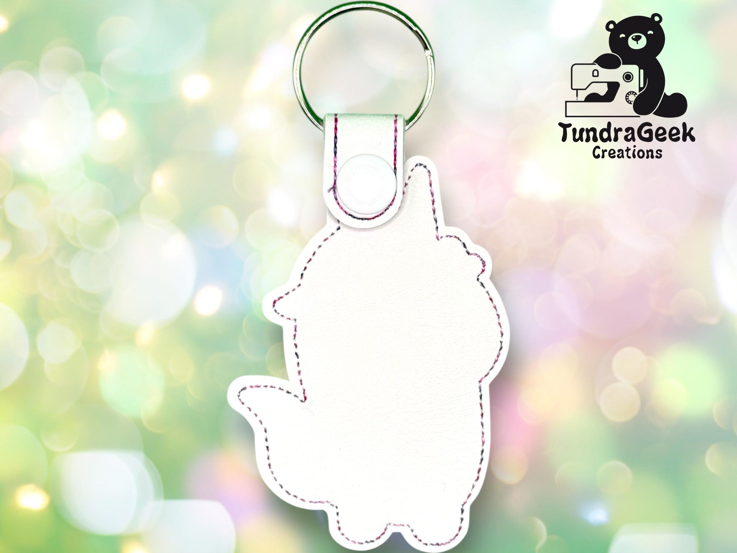 A white, unicorn-shaped keychain with pink stitching is shown against a blurred, colorful background. The TundraGeek Creations logo appears in the top right corner.