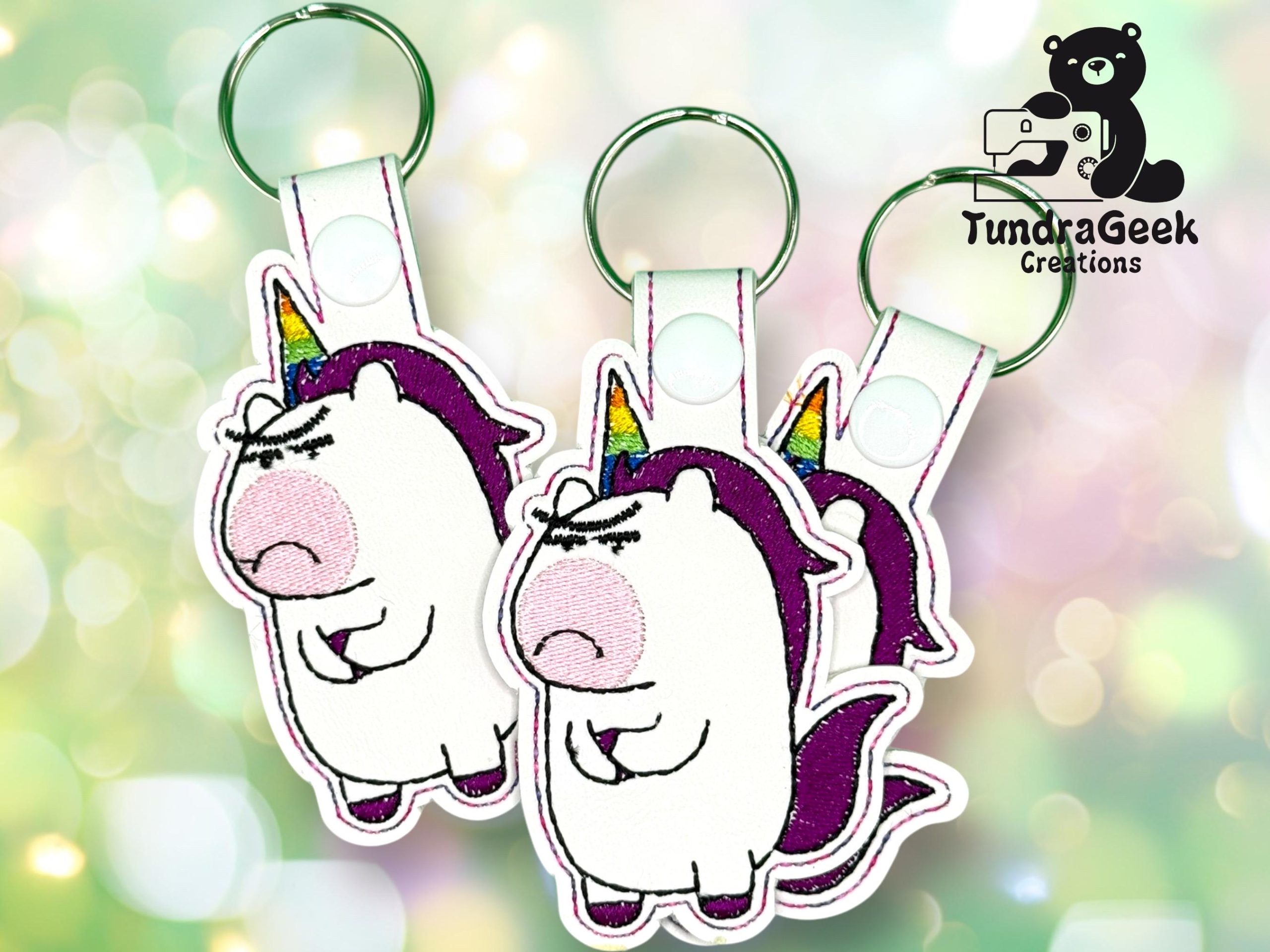 Three embroidered keychains with an angry unicorn design, featuring a rainbow horn and purple mane, displayed against a light, bokeh background. TundraGeek Creations logo in the corner.