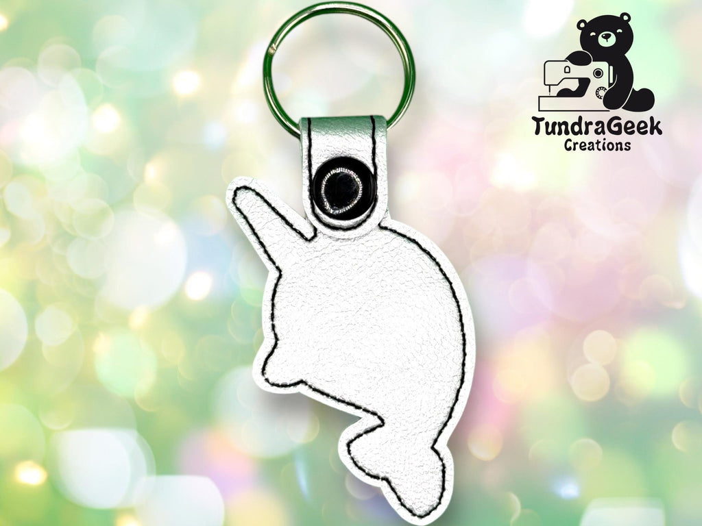 A white stitched keychain in the shape of a narwhal with a green keyring, displayed on a blurred green background with the TundraGeek Creations logo in the corner.