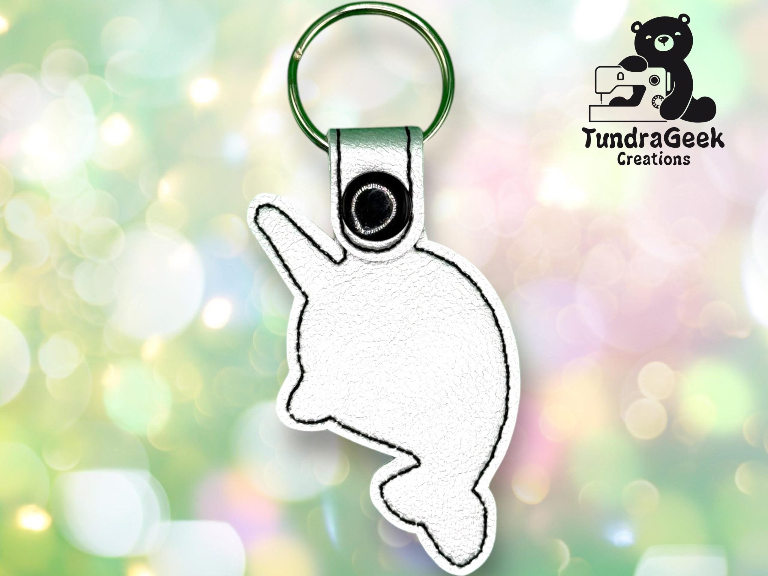 A white stitched keychain in the shape of a narwhal with a green keyring, displayed on a blurred green background with the TundraGeek Creations logo in the corner.