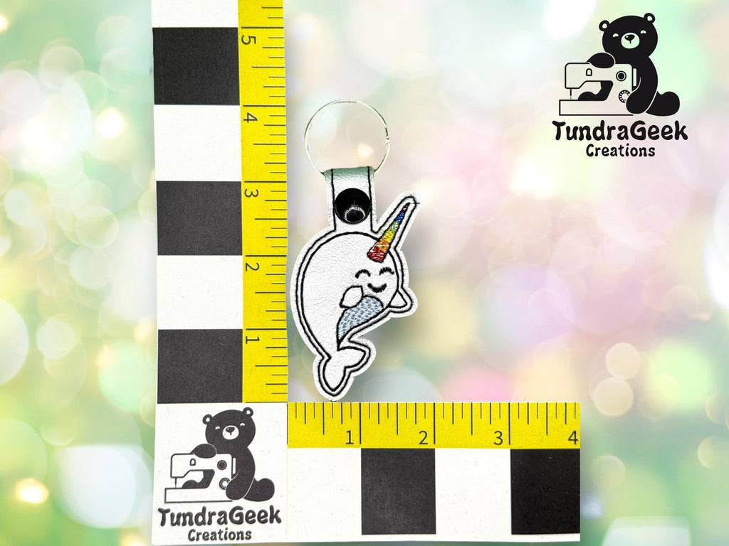 A keychain featuring an embroidered narwhal with a rainbow horn is placed next to a ruler for scale. The TundraGeek Creations logo is visible in the image.