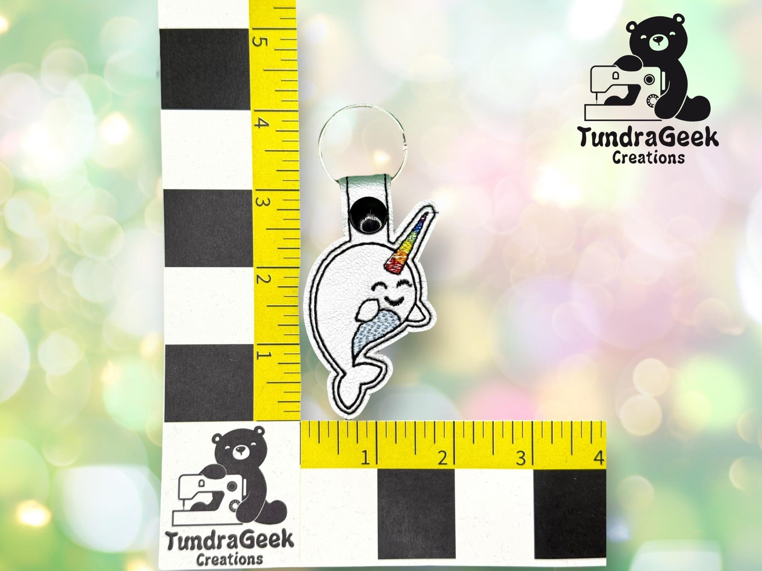 A keychain featuring an embroidered narwhal with a rainbow horn is placed next to a ruler for scale. The TundraGeek Creations logo is visible in the image.