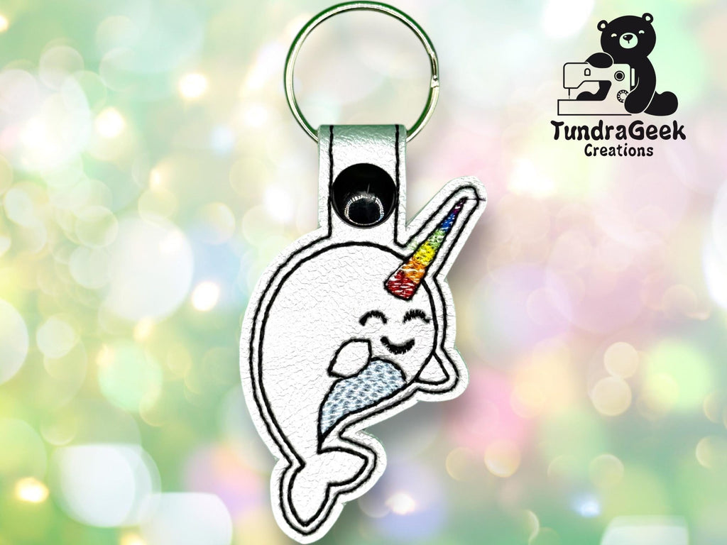 Keychain featuring an embroidered cartoon narwhal with a rainbow horn, set against a soft-focus, colorful background. TundraGeek Creations logo appears in the top right corner.