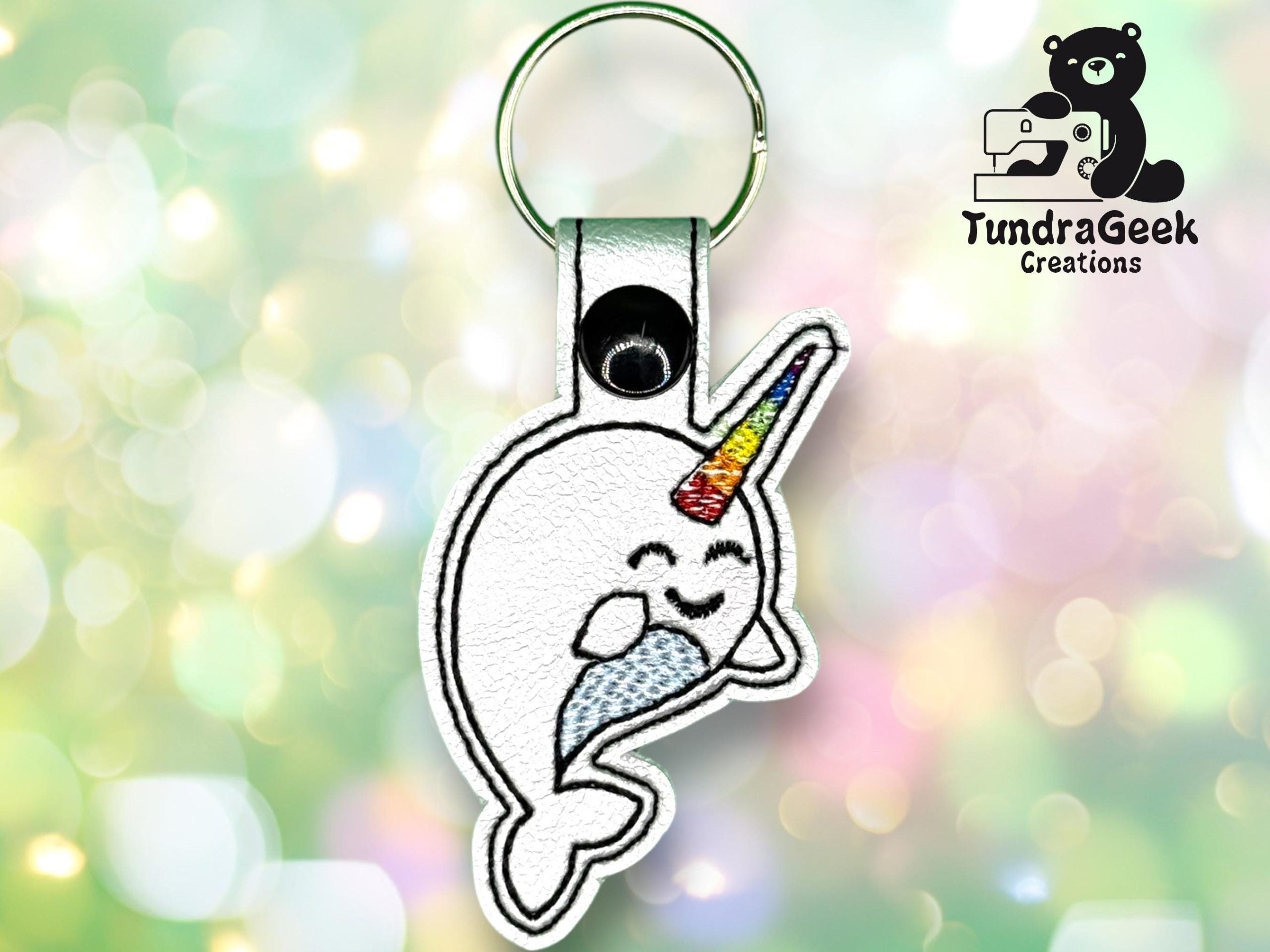 Keychain featuring an embroidered cartoon narwhal with a rainbow horn, set against a soft-focus, colorful background. TundraGeek Creations logo appears in the top right corner.