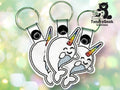 Three white narwhal keychains with rainbow-colored horns and closed eyes are displayed against a blurred, light background. A Tundra Geek Creations logo featuring a bear is in the top right corner.