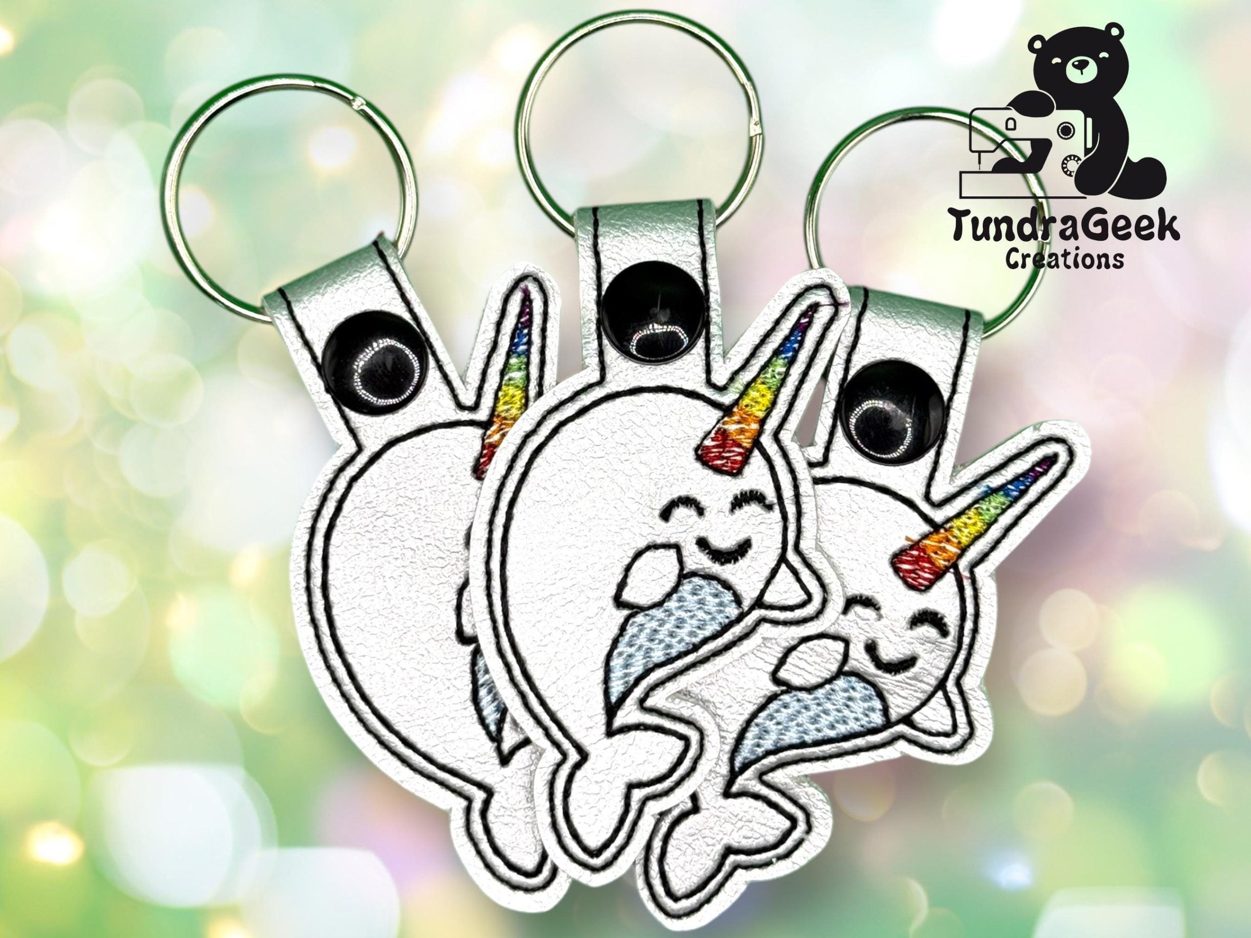 Three white narwhal keychains with rainbow-colored horns and closed eyes are displayed against a blurred, light background. A Tundra Geek Creations logo featuring a bear is in the top right corner.