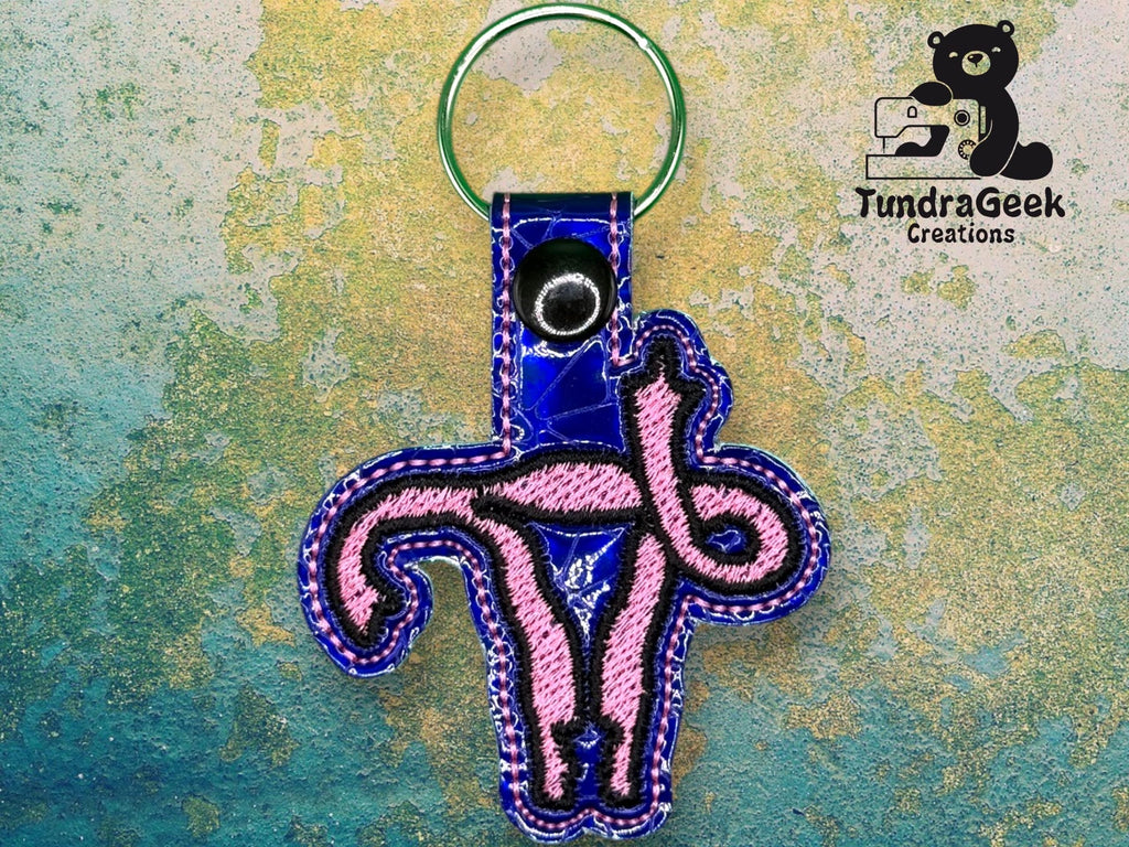 A keychain shaped like a pink and blue uterus with a snap button and key ring, placed on a textured background. TundraGeek Creations logo is in the top right corner.