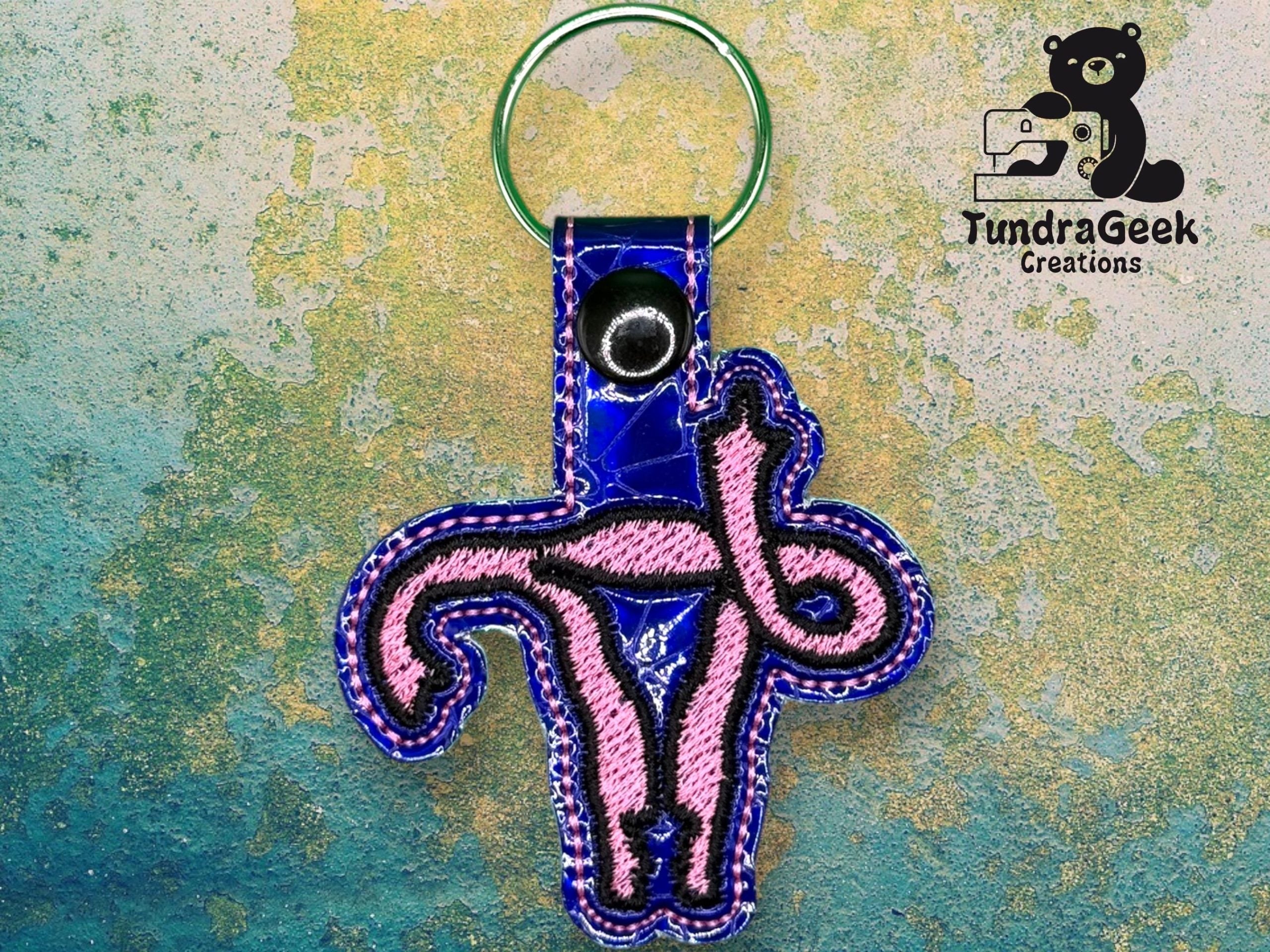 A keychain with a pink embroidered uterus design on blue fabric, featuring a green keyring and a silver snap, displayed on a textured background with the TundraGeek Creations logo.