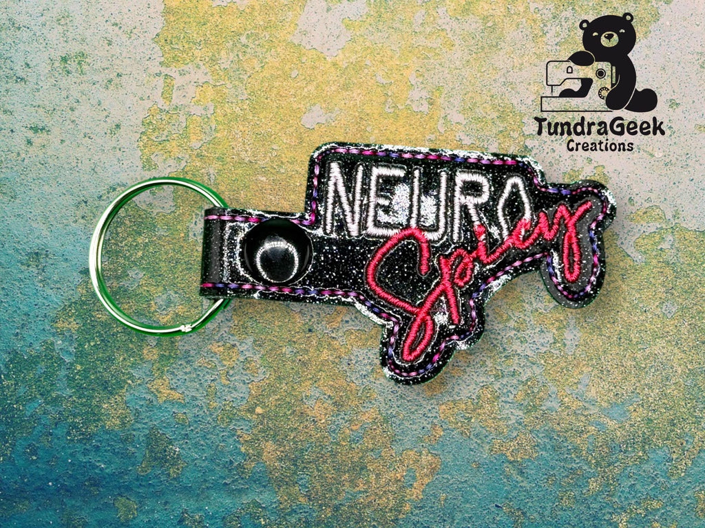 Keychain with embroidered text NEURO Spicy in black, white, and red thread, next to the TundraGeek Creations logo featuring a bear with a sewing machine.