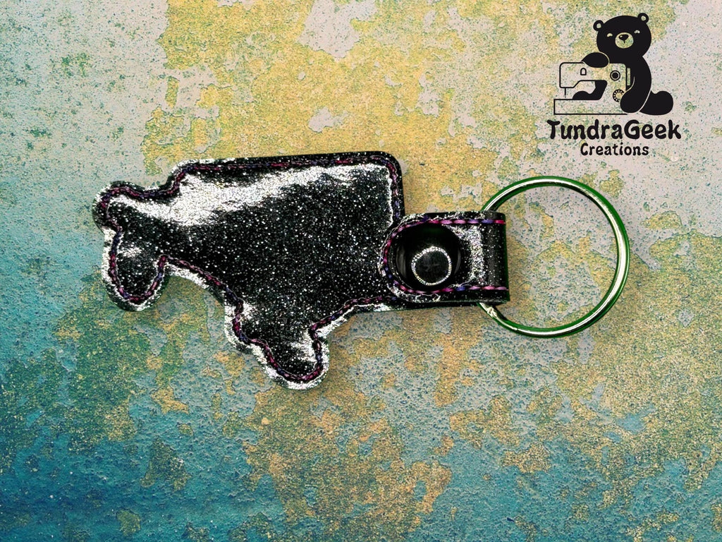 A metallic keychain shaped like a bison with a keyring, placed on a textured background. TundraGeek Creations logo with a bear appears in the top right corner.