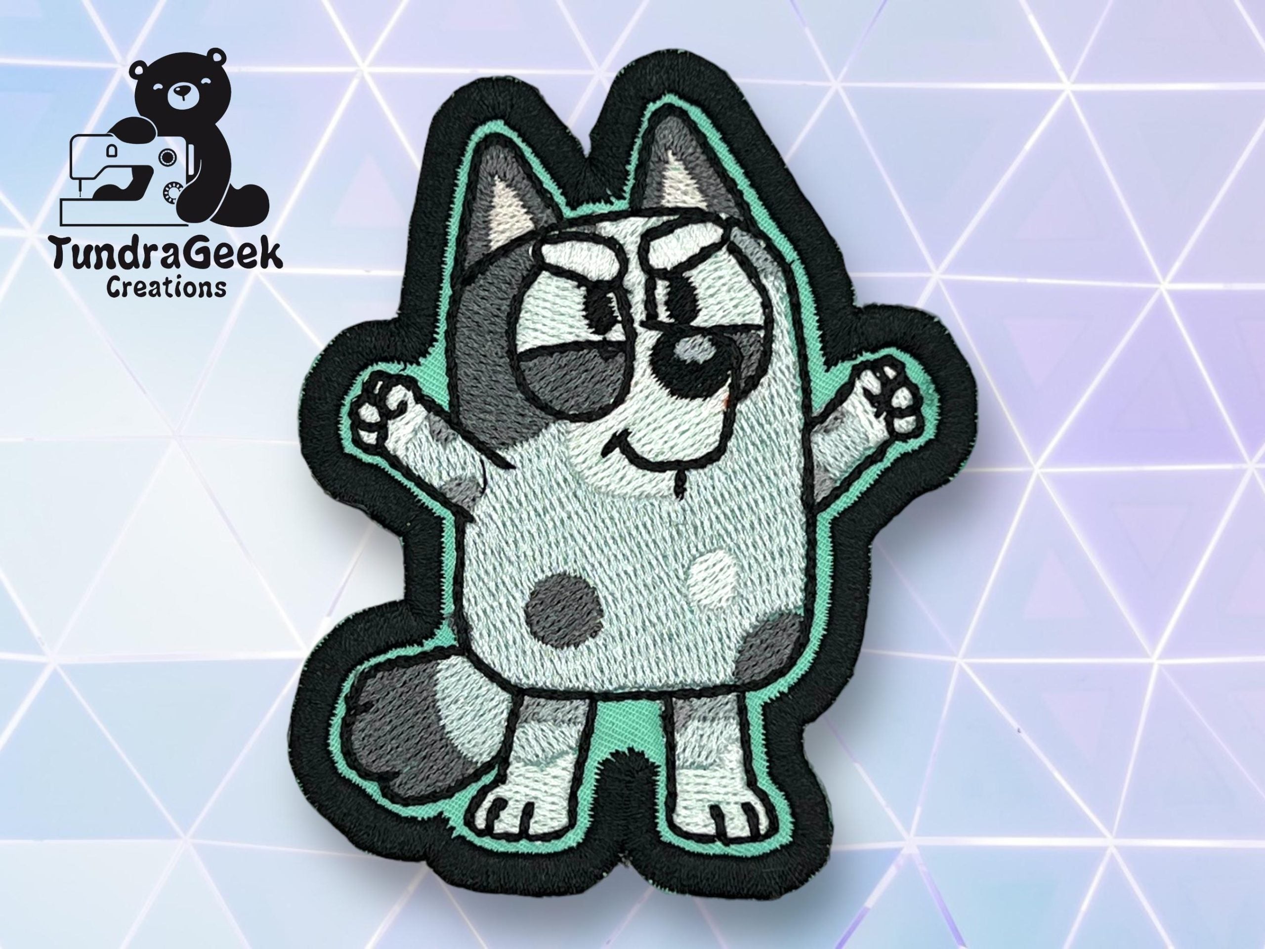An embroidered patch of a cartoon gray and white dog with raised arms, black outlines, and a confident expression. TundraGeek Creations logo is in the top left corner.
