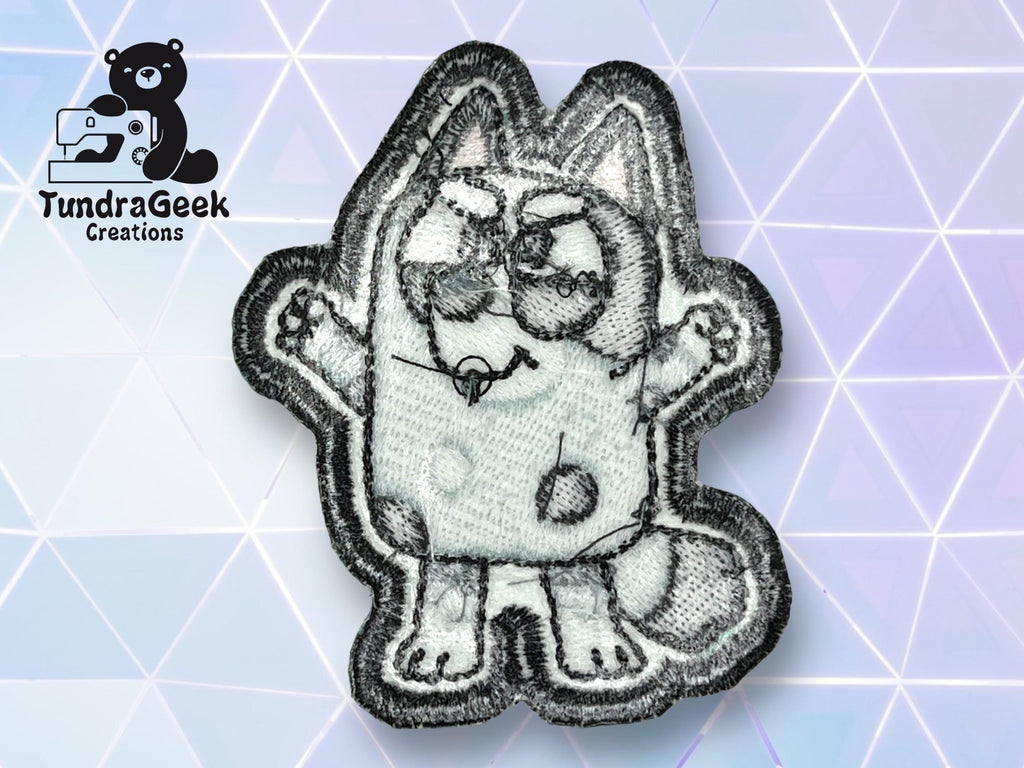 An embroidered patch featuring a cartoon cat with glasses, raised arms, and spots, displayed on a geometric, pastel background. TundraGeek Creations logo is in the top left corner.