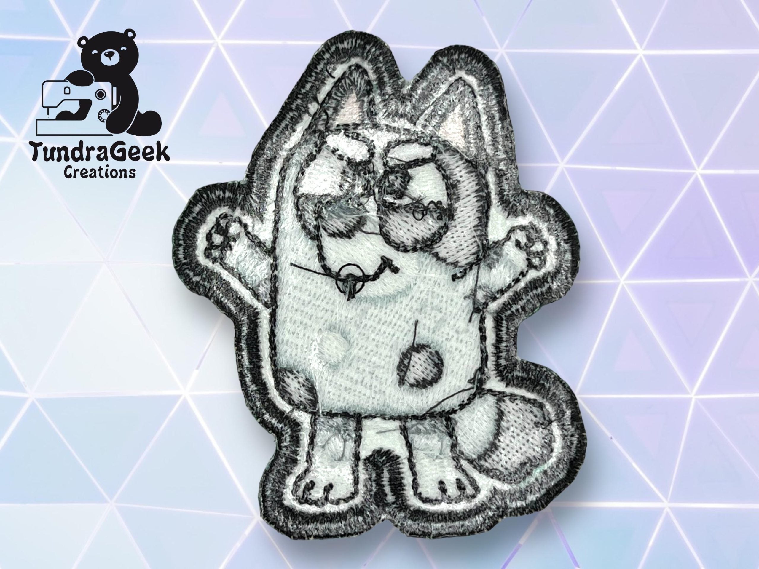 An embroidered patch featuring a cartoon cat with glasses, raised arms, and spots, displayed on a geometric, pastel background. TundraGeek Creations logo is in the top left corner.