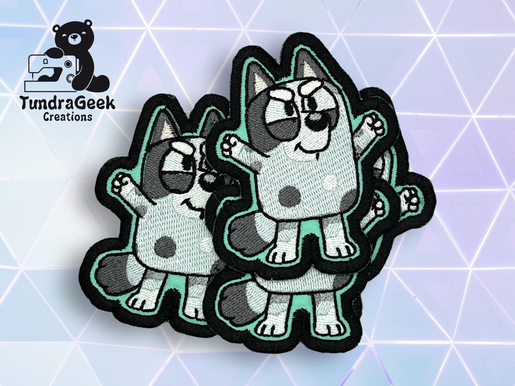 Three embroidered patches featuring a cartoon blue dog with spots and raised arms, displayed on a geometric light background. TundraGeek Creations logo in the top left corner.