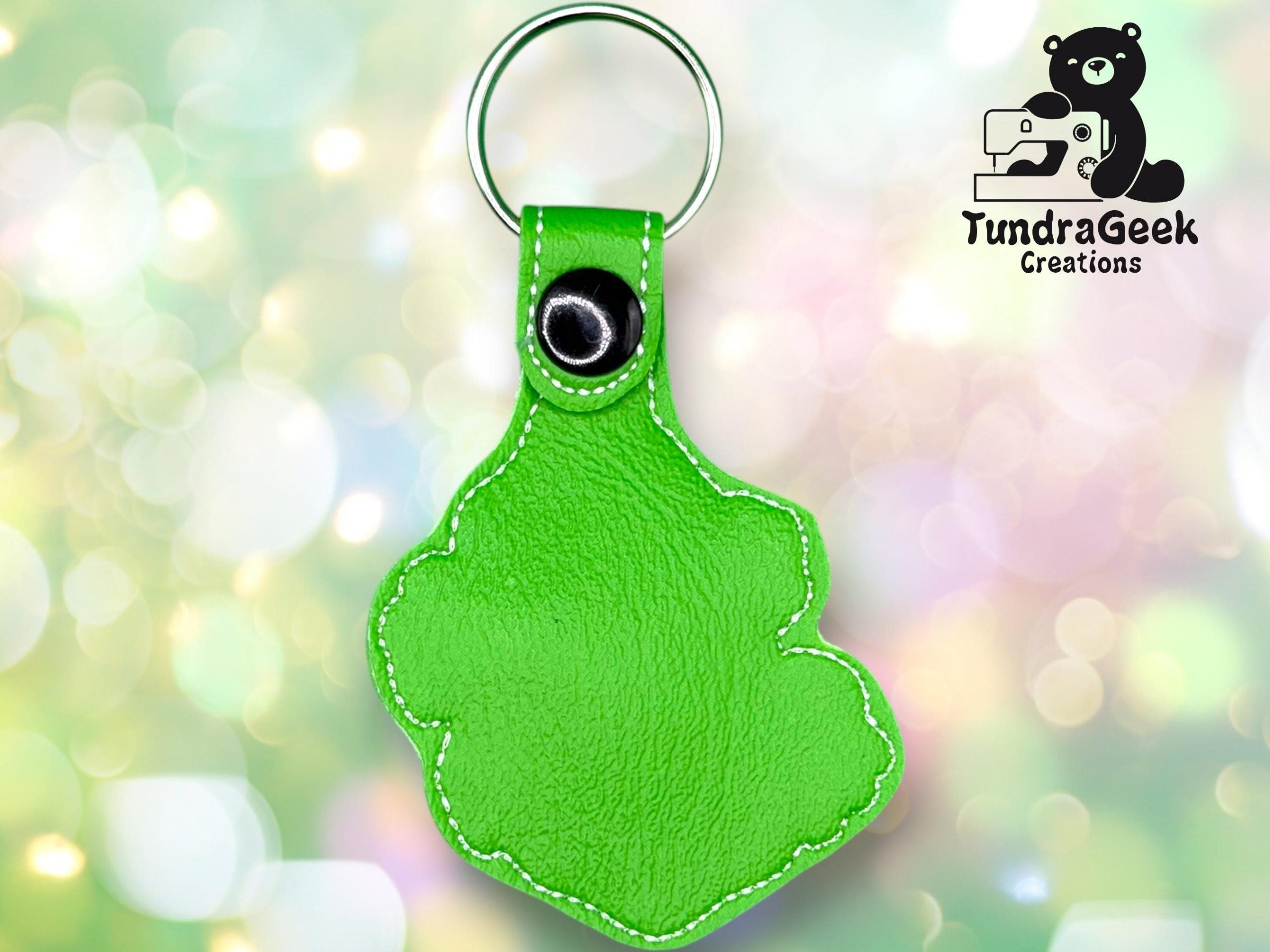Bright green, paw-shaped keychain with white stitching and metal ring, displayed against a blurred pastel background. TundraGeek Creations logo with bear and sewing machine in top right corner.