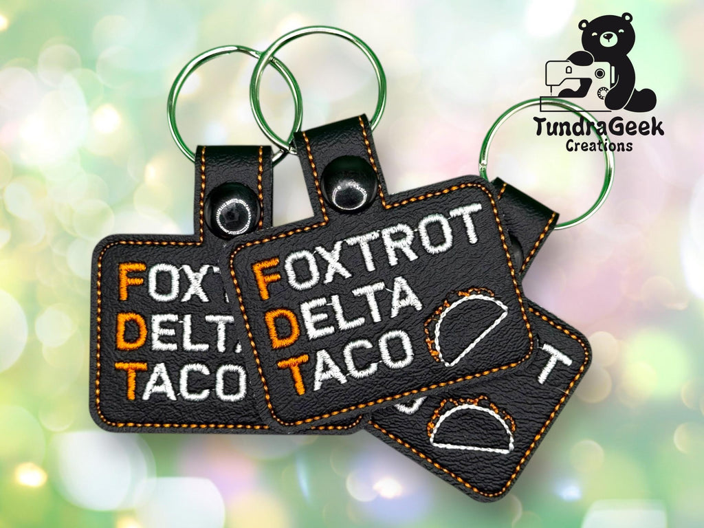 Three black keychains with orange stitching display the words Foxtrot Delta Taco in white and orange, with a taco graphic and a Tundra Geek Creations logo in the background.
