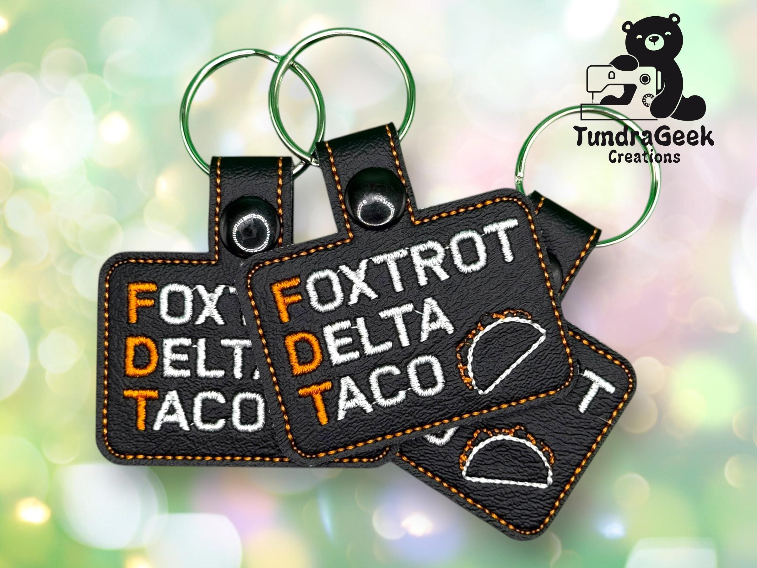 Three black keychains with orange stitching display the words Foxtrot Delta Taco in white and orange, with a taco graphic and a Tundra Geek Creations logo in the background.