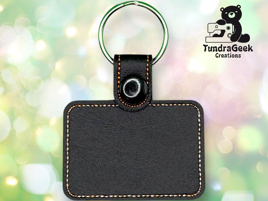 Rectangular black leather keychain with contrast stitching and a metal ring, displayed against a blurred, colorful background with the TundraGeek Creations logo in the upper right corner.