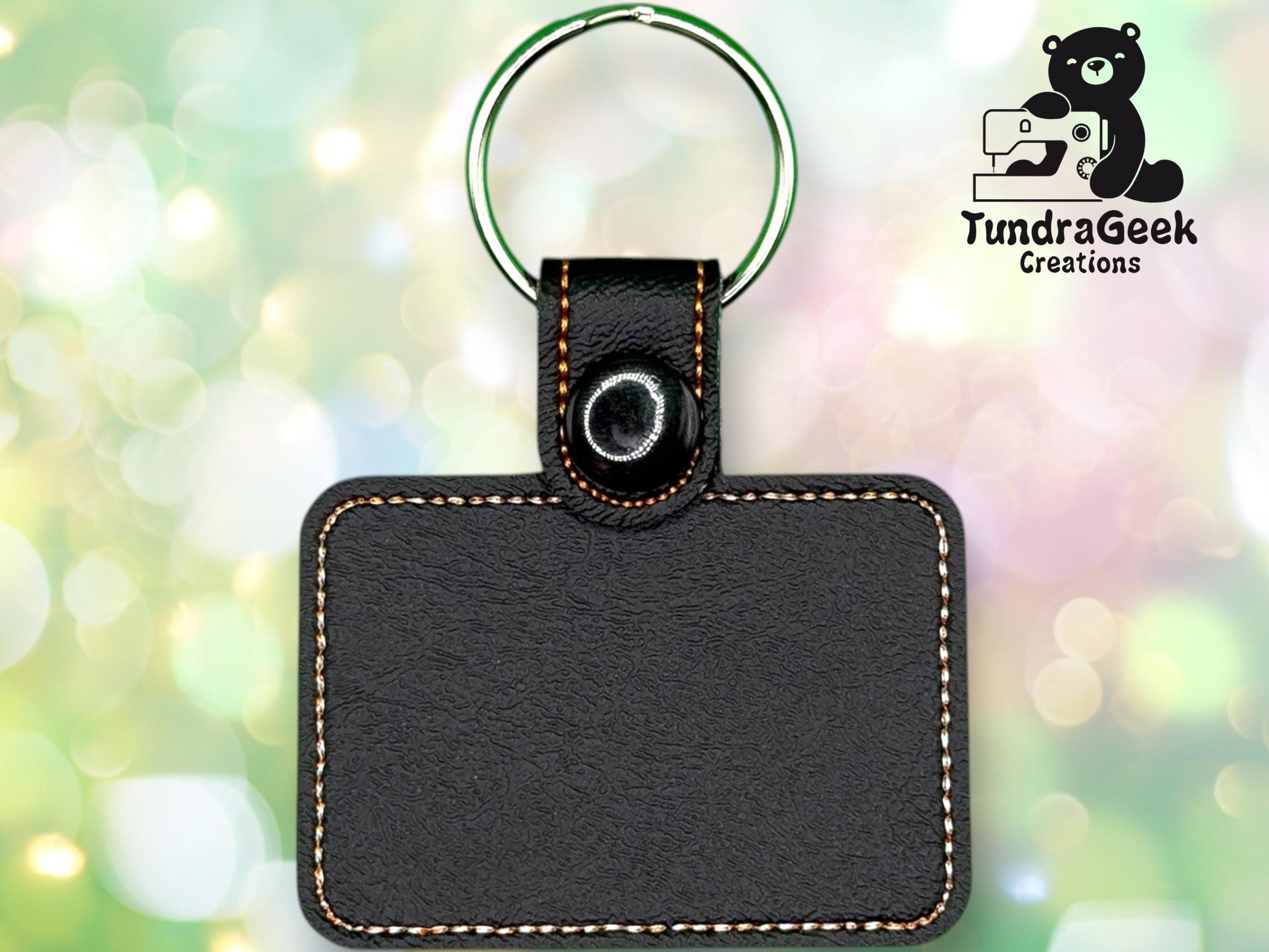 Rectangular black leather keychain with contrast stitching and a metal ring, displayed against a blurred, colorful background with the TundraGeek Creations logo in the upper right corner.