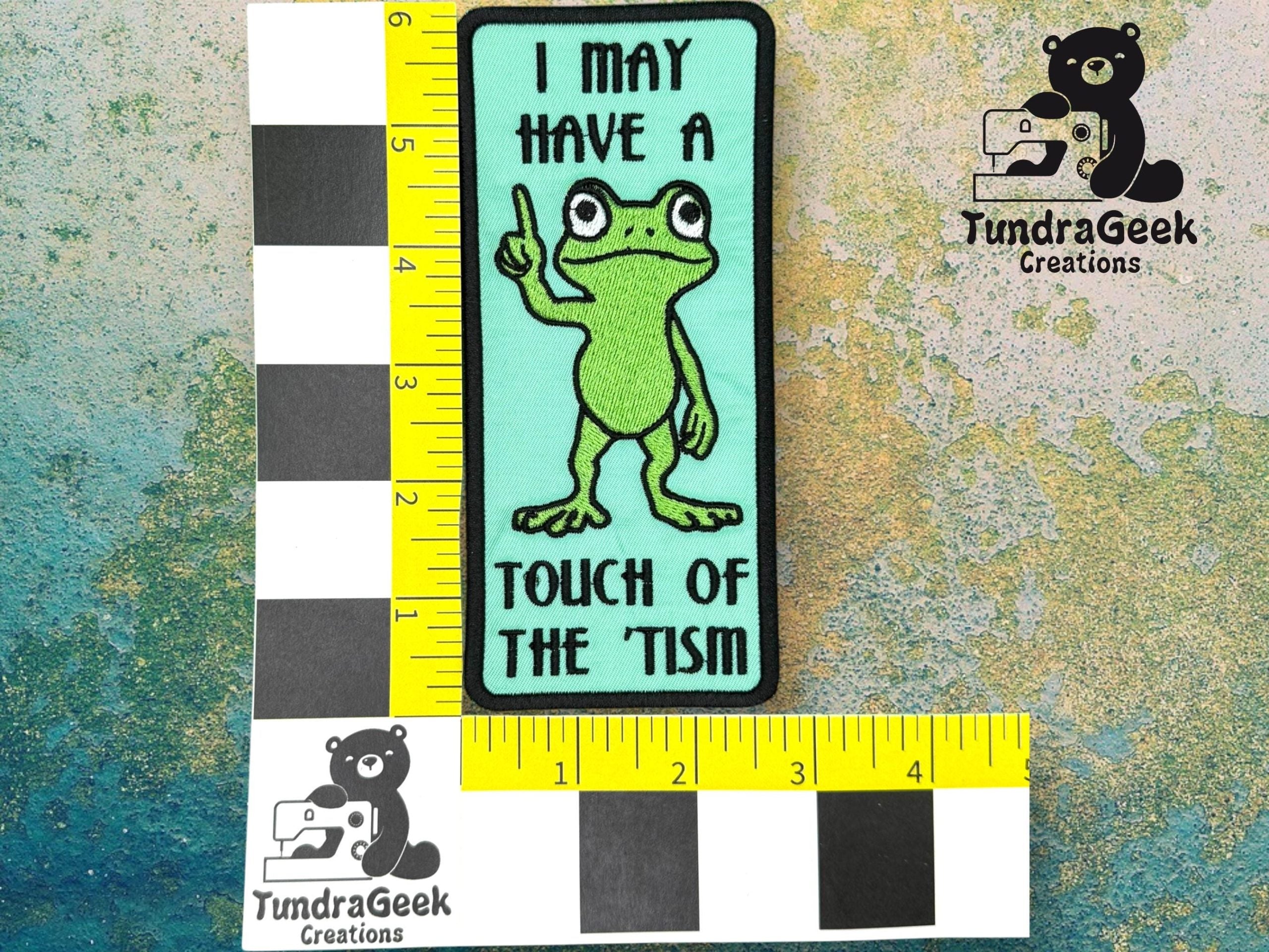 A rectangular patch with a cartoon frog raising one finger and text that reads, I may have a touch of the tism. Measuring tape and TundraGeek Creations logo are visible.