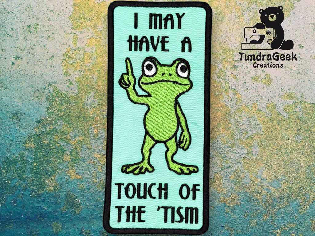 Embroidered patch with a cartoon frog holding up one finger; text reads “I may have a touch of the tism.” TundraGeek Creations logo is in the corner.