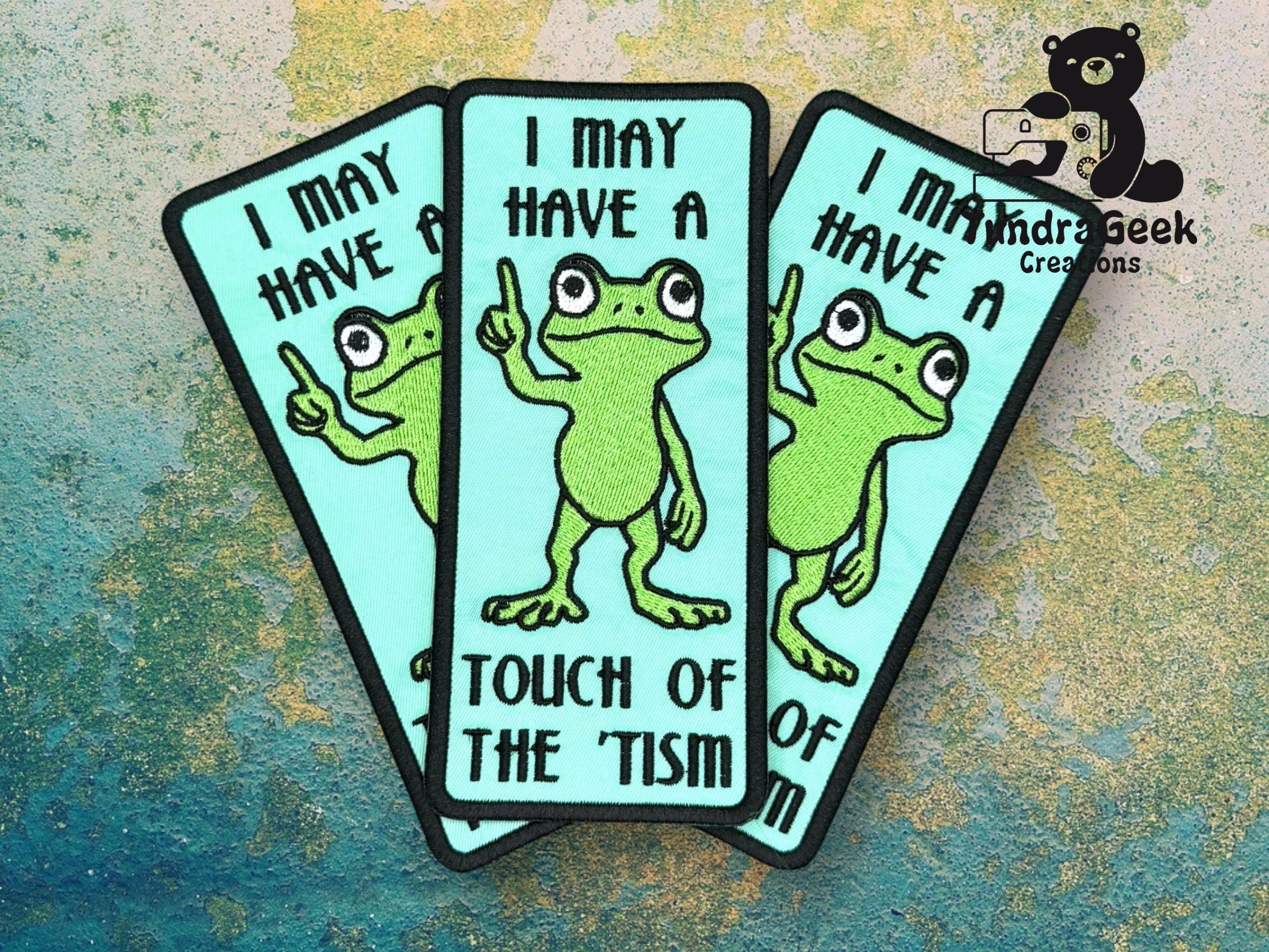 Three identical patches show a cartoon frog pointing upward with the text, I may have a touch of the tism. Background has a blue and green textured pattern.