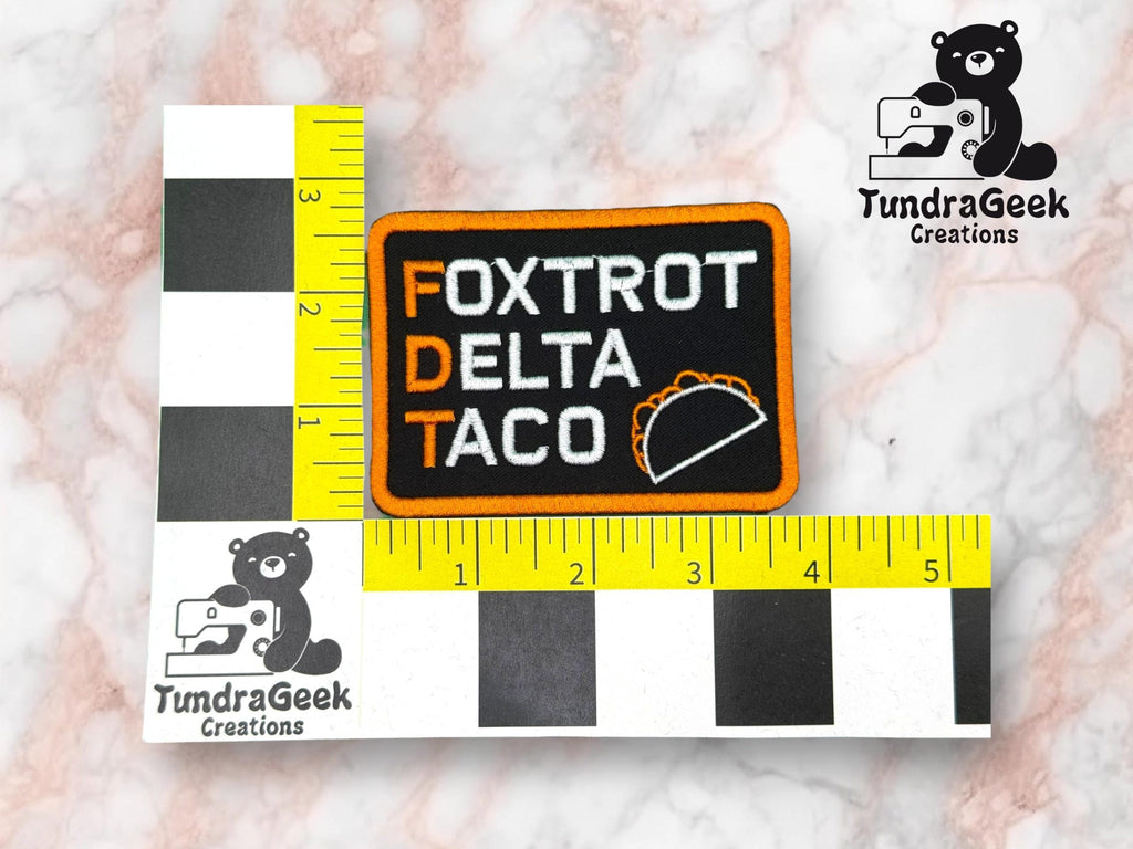 A rectangular embroidered patch labeled Foxtrot Delta Taco with orange border and taco graphic, placed next to a black-and-white inch ruler for scale.