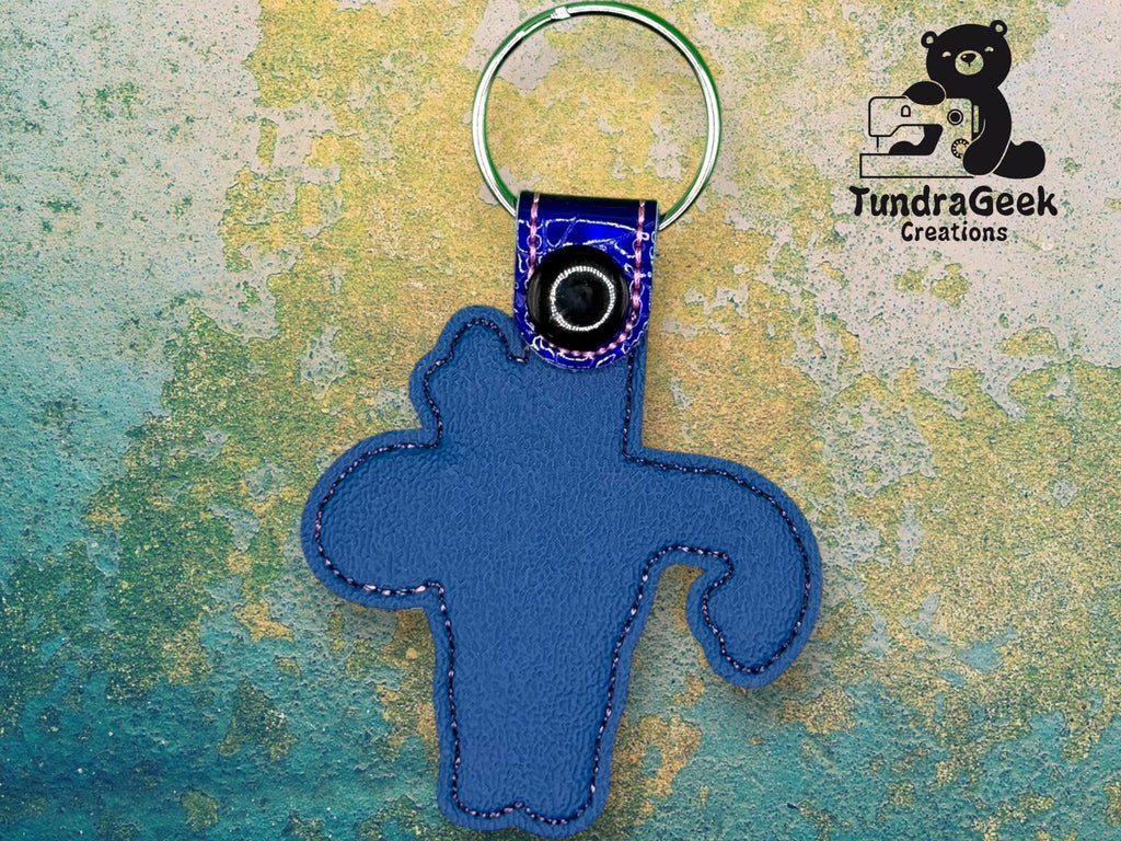 Blue cactus-shaped keychain with a metal ring, placed on a textured background. TundraGeek Creations logo featuring a bear with a sewing machine is in the upper right corner.