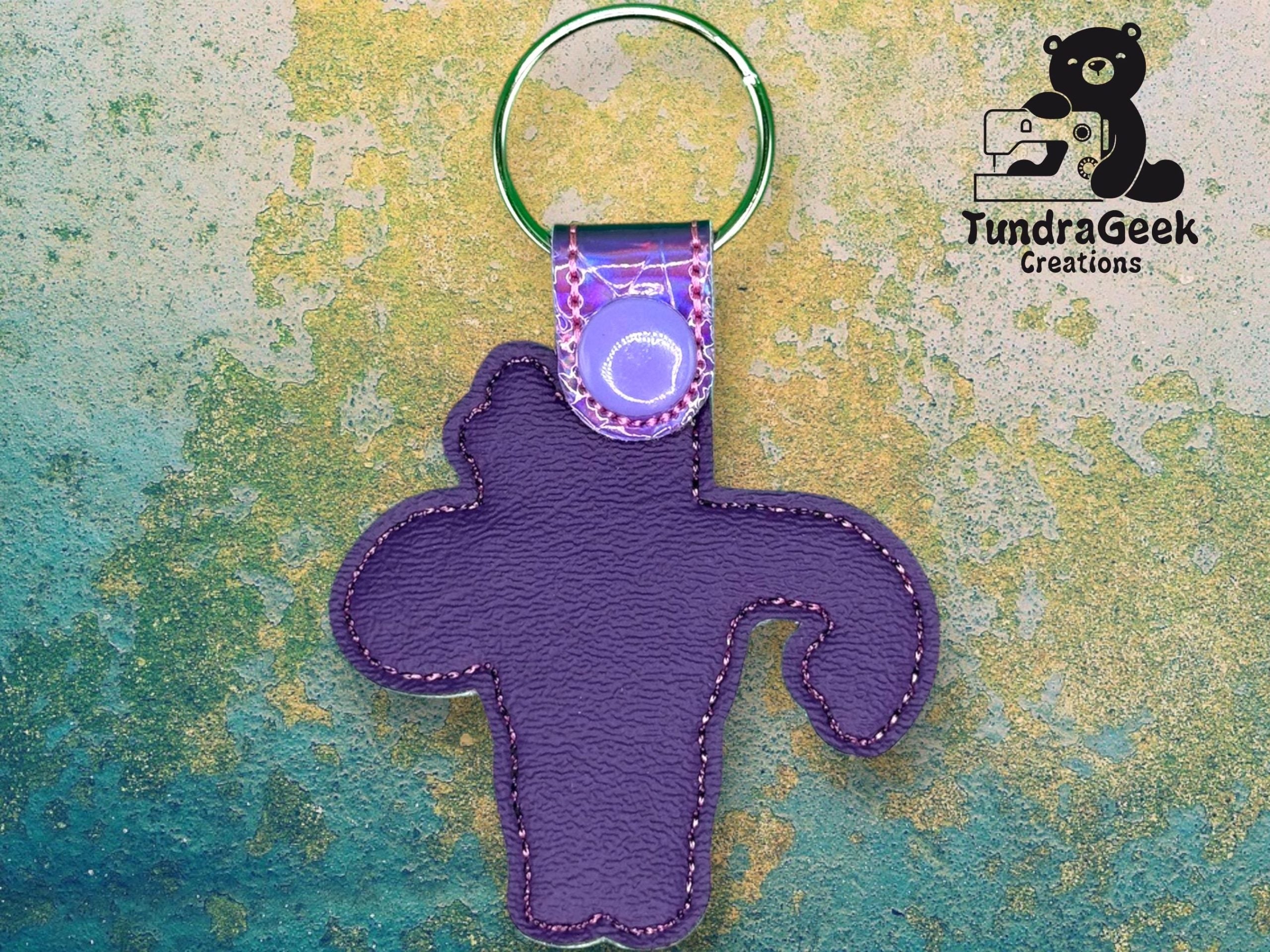 A purple leather keychain shaped like a bear, attached to a metal ring, displayed against a textured background with the TundraGeek Creations logo in the corner.