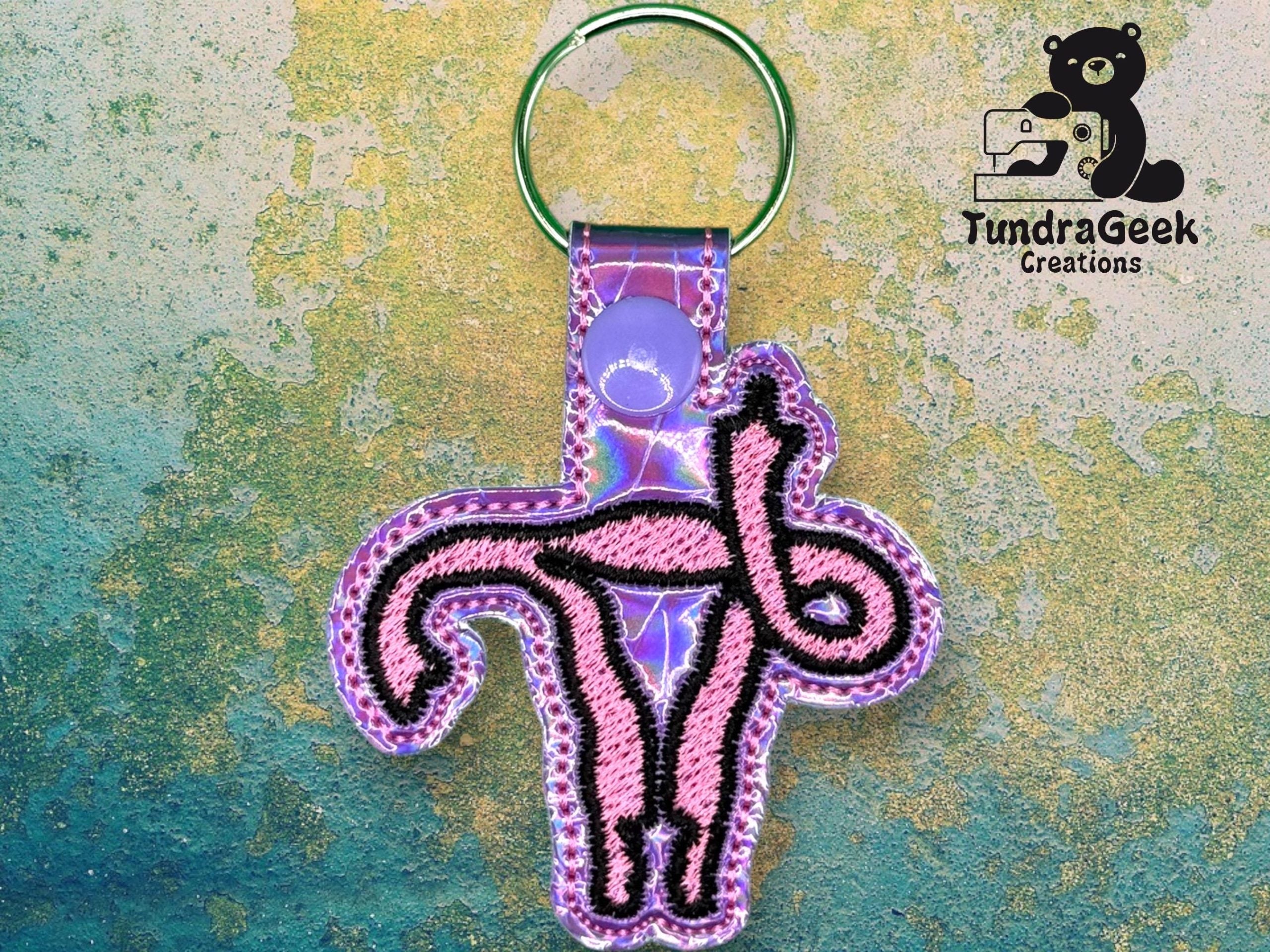 A keychain featuring an embroidered purple design of the Nazca Lines monkey geoglyph, with a green keyring and the TundraGeek Creations logo in the top right corner.
