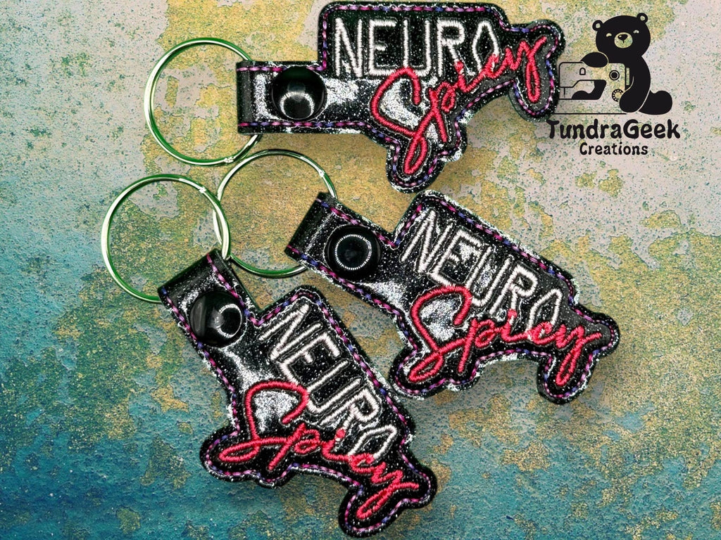 Three glittery keychains with the words Neuro Spicy in bold and script fonts, displayed on a textured blue and green surface. TundraGeek Creations logo in the top right corner.