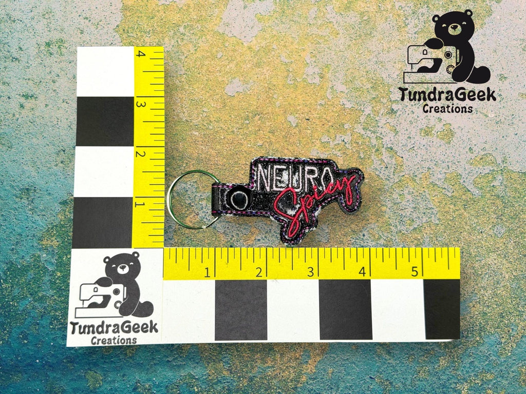 A keychain reading Neuro Spicy is displayed on a measurement grid, showing it is about 3 inches long. The TundraGeek Creations logo appears twice on the image.