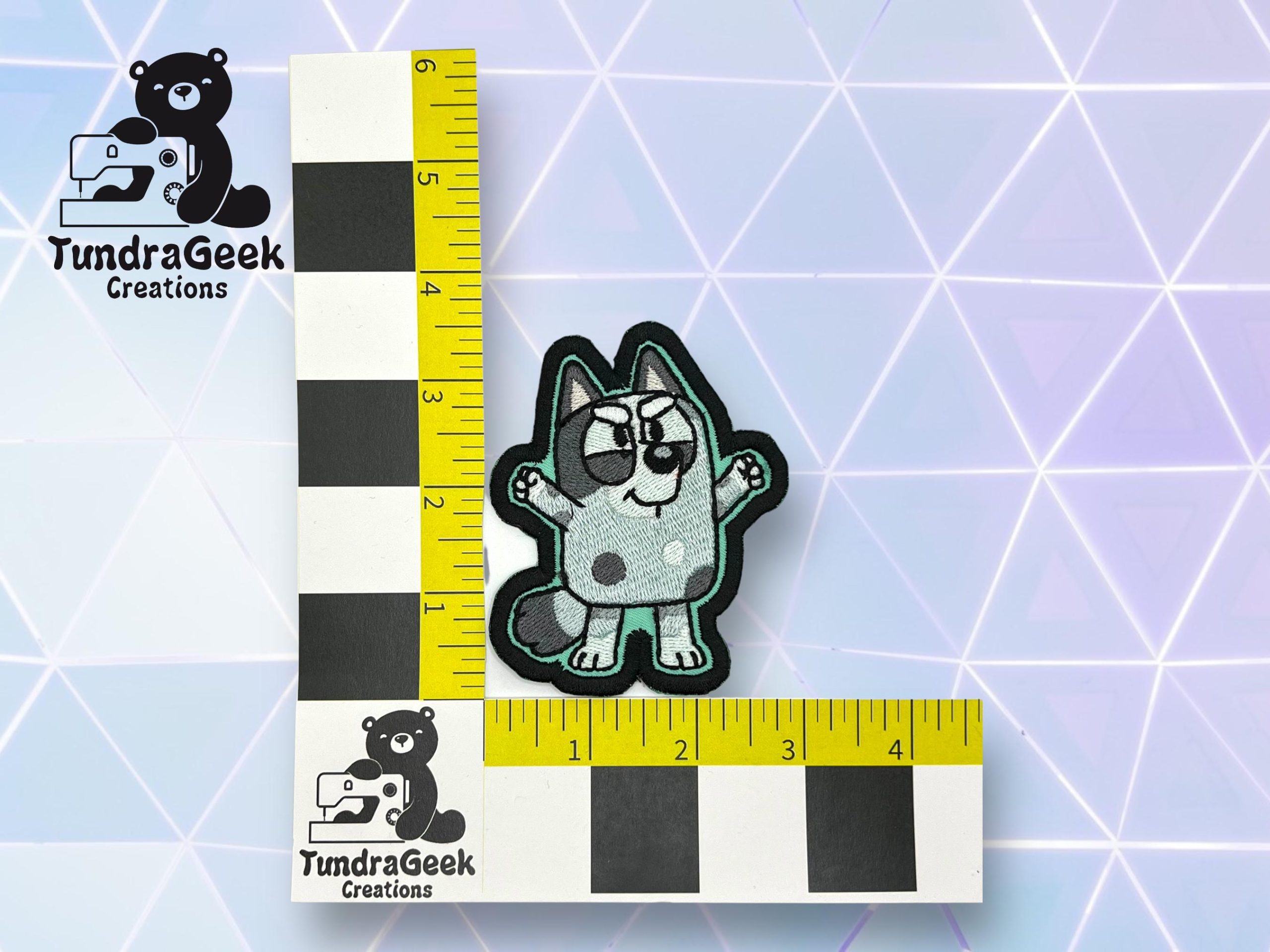 A small embroidered patch of a cartoon dog is placed on a checkered measuring grid with TundraGeek Creations logos on a geometric background.
