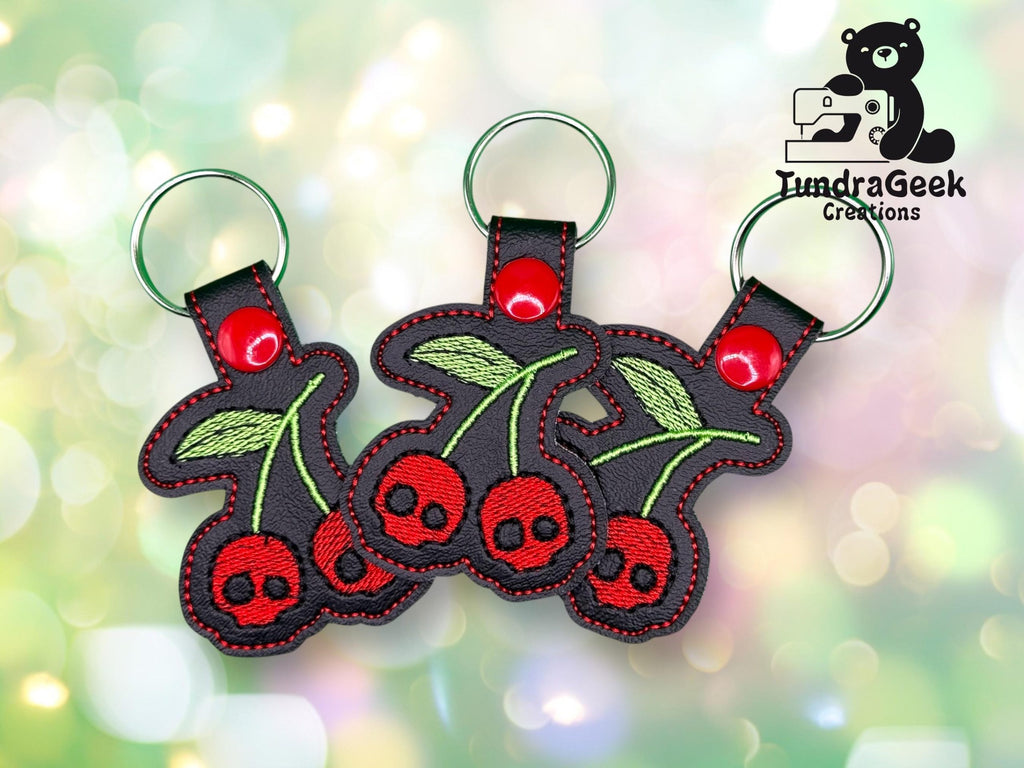 Three embroidered cherry keychains with red skull-shaped cherries and green leaves, shown on a blurred, colorful background. TundraGeek Creations logo with a bear and sewing machine in the corner.