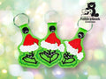 Three green keychains depicting a cartoon character in a Santa hat forming a heart with its hands, displayed against a light bokeh background. TundraGeek Creations logo in the top right corner.
