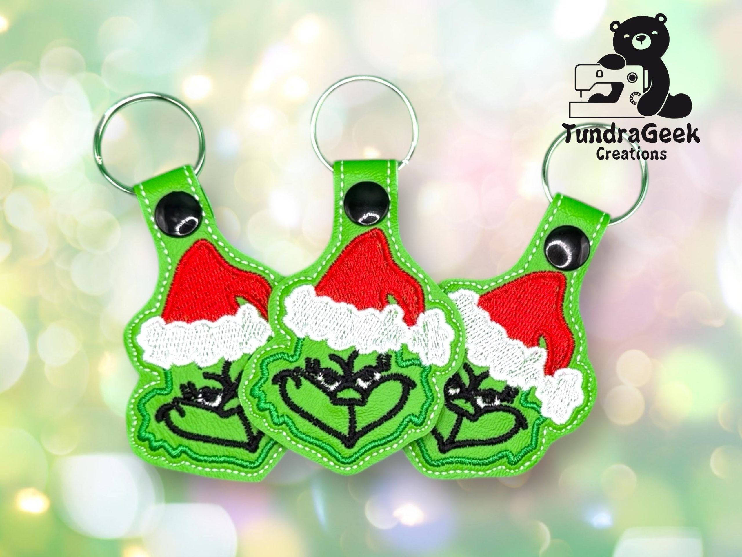 Three green keychains depicting a cartoon character in a Santa hat forming a heart with its hands, displayed against a light bokeh background. TundraGeek Creations logo in the top right corner.