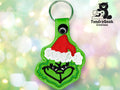 A green Grinch face keychain with a red Santa hat and black outline, displayed against a light bokeh background. Logo in the corner reads TundraGeek Creations with a bear and sewing machine.
