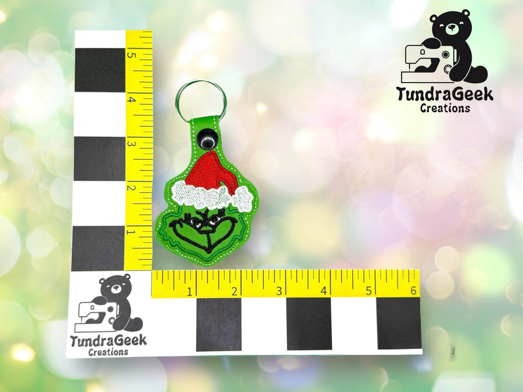A green keychain with a red Santa hat design is placed next to a ruler for scale, with the TundraGeek Creations logo in the background.
