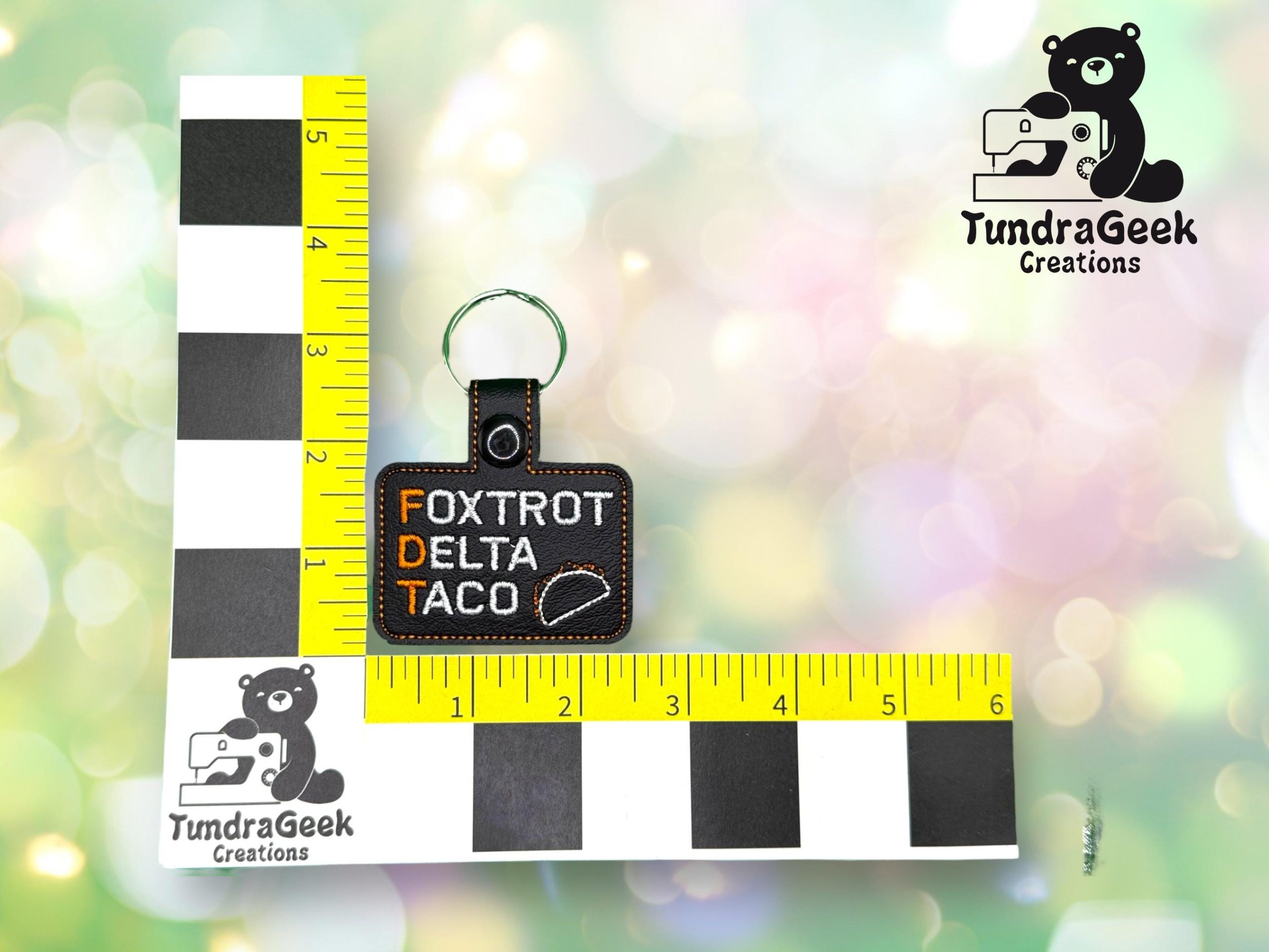 A rectangular keychain reads Foxtrot Delta Taco with a taco icon, placed on an L-shaped ruler marked in inches. TundraGeek Creations logo is visible.