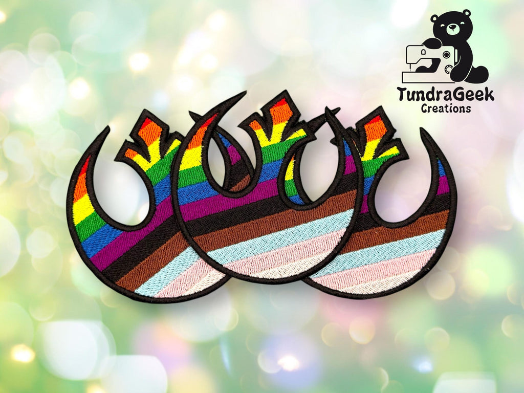 Three embroidered patches featuring the Star Wars Rebel Alliance symbol with rainbow stripes, representing LGBTQ+ pride flags, on a blurred green background with a TundraGeek Creations logo.