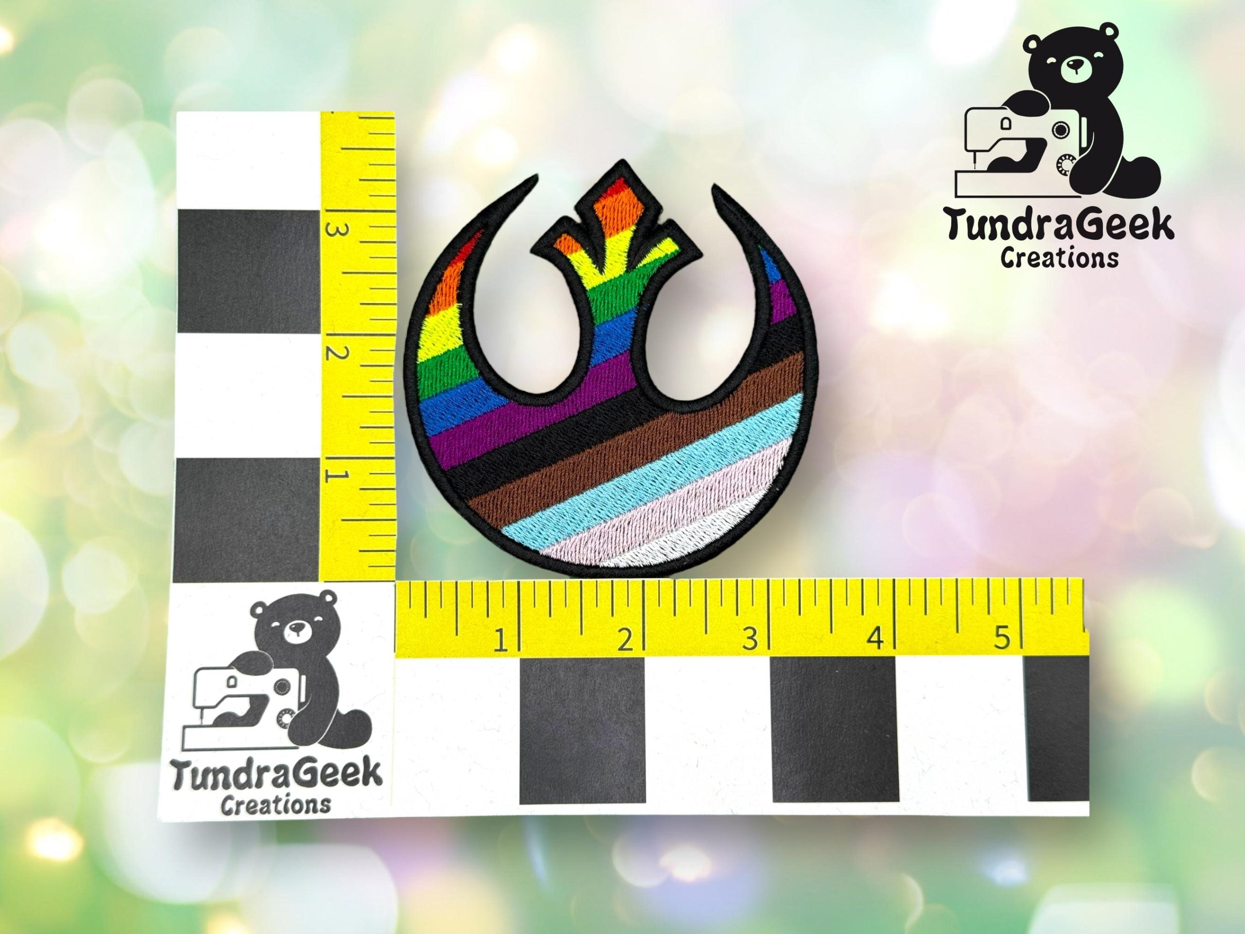 A patch with a rainbow-striped symbol is positioned on a measuring grid showing it is about 3 inches wide. The TundraGeek Creations logo appears in two corners.