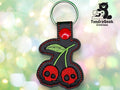 Keychain with two red cherries embroidered as skulls on black faux leather, with a red snap and green stitching. TundraGeek Creations logo is in the top right corner.