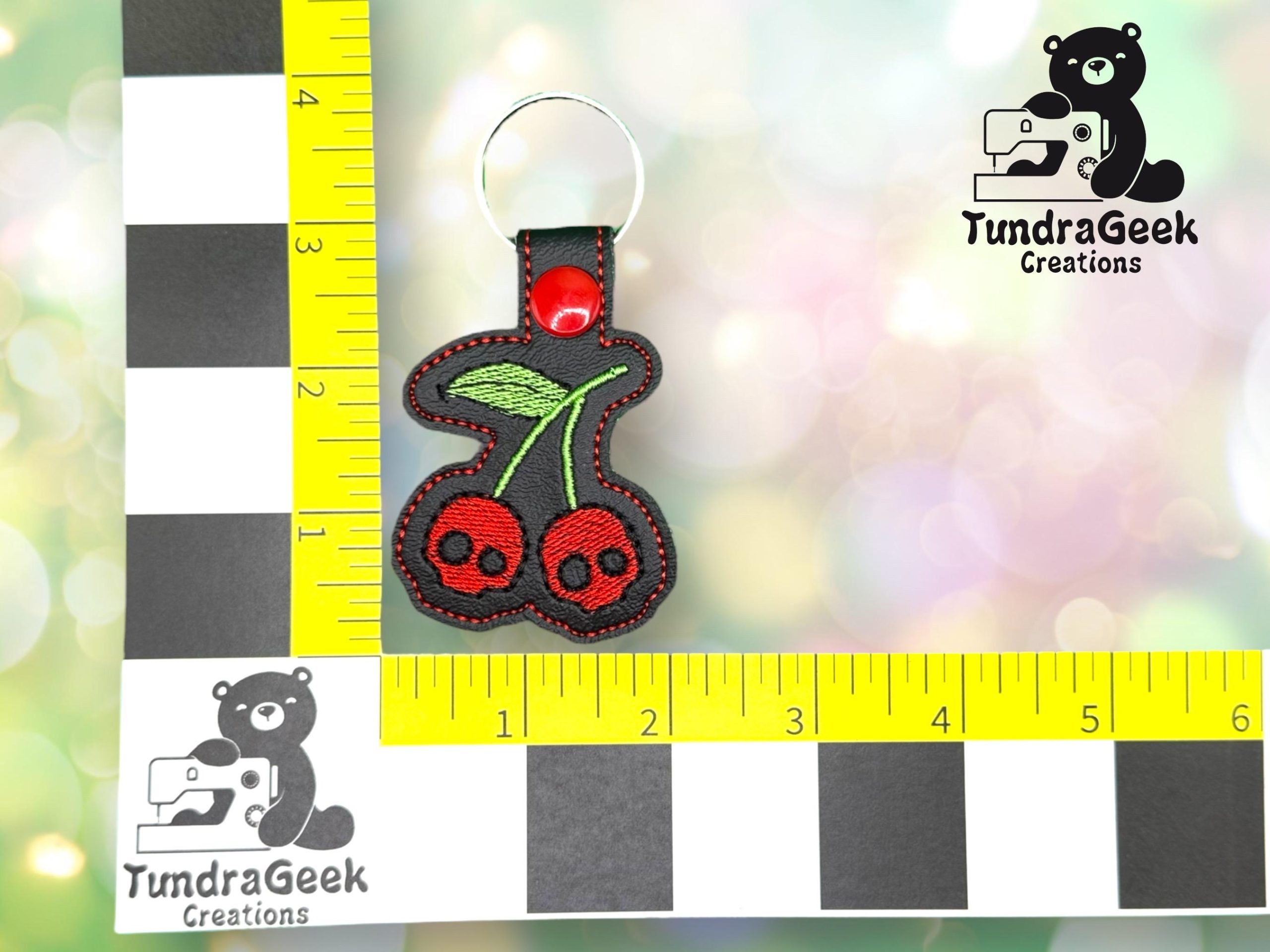 Embroidered keychain with two red cherries featuring black skulls, placed on a measuring mat with TundraGeek Creations logos.